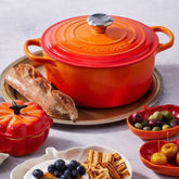 Panela redonda de ferro fundido Le Creuset laranja em mesa posta com pão, petisqueiras com azeitonas e frutas, ideal para receitas sofisticadas. Combina charme, durabilidade e alta performance na cozinha.